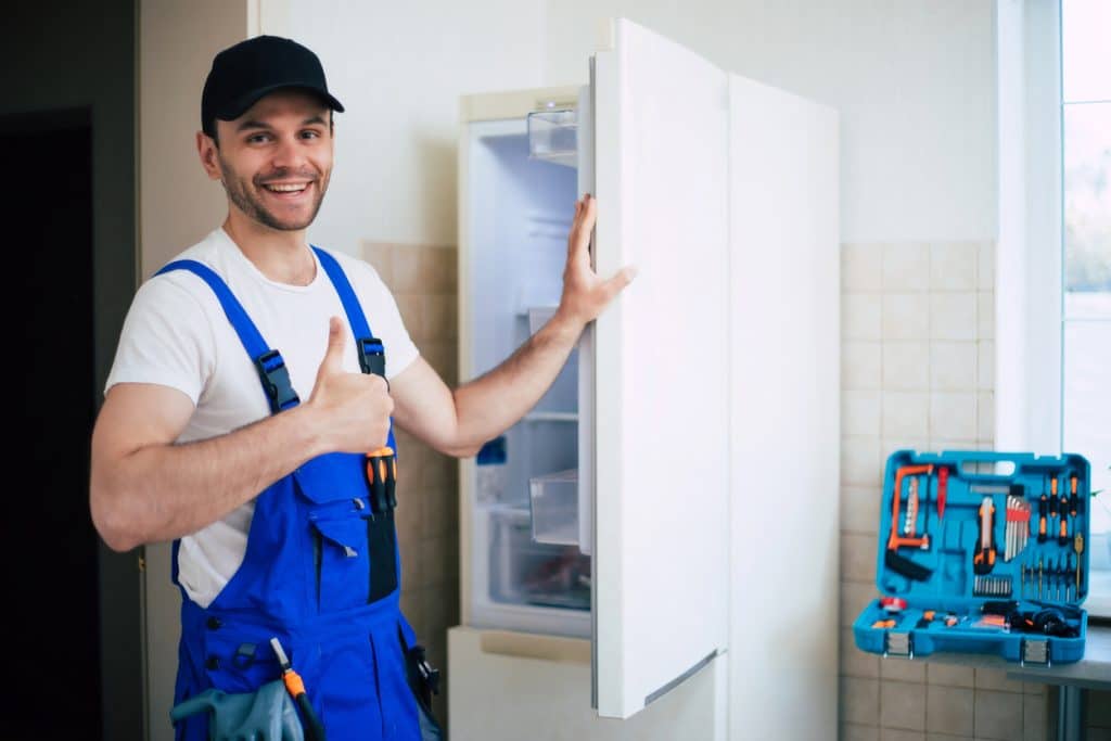 Professional young repairman in worker uniform and cap with modern toolbox with equipment after repair of refrigerator on the kitchen